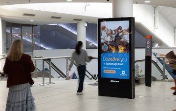 An indoor public walkway with tiled floors and glass railings features a freestanding digital advertising display. 